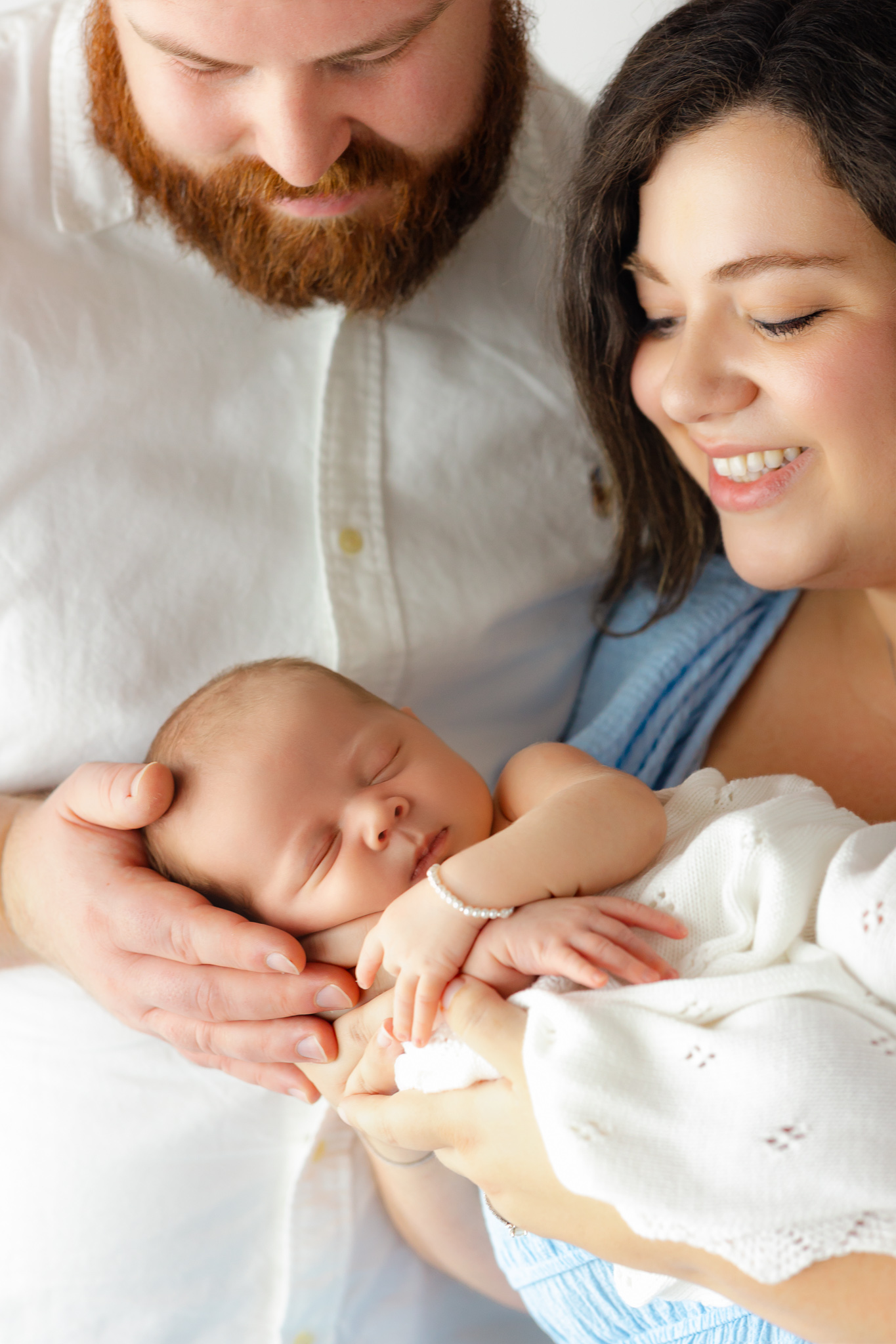 A newborn baby sleeps in a white blanket in mom and dad's hands as they smile down to her after getting placenta encapsulation in Savannah, GA