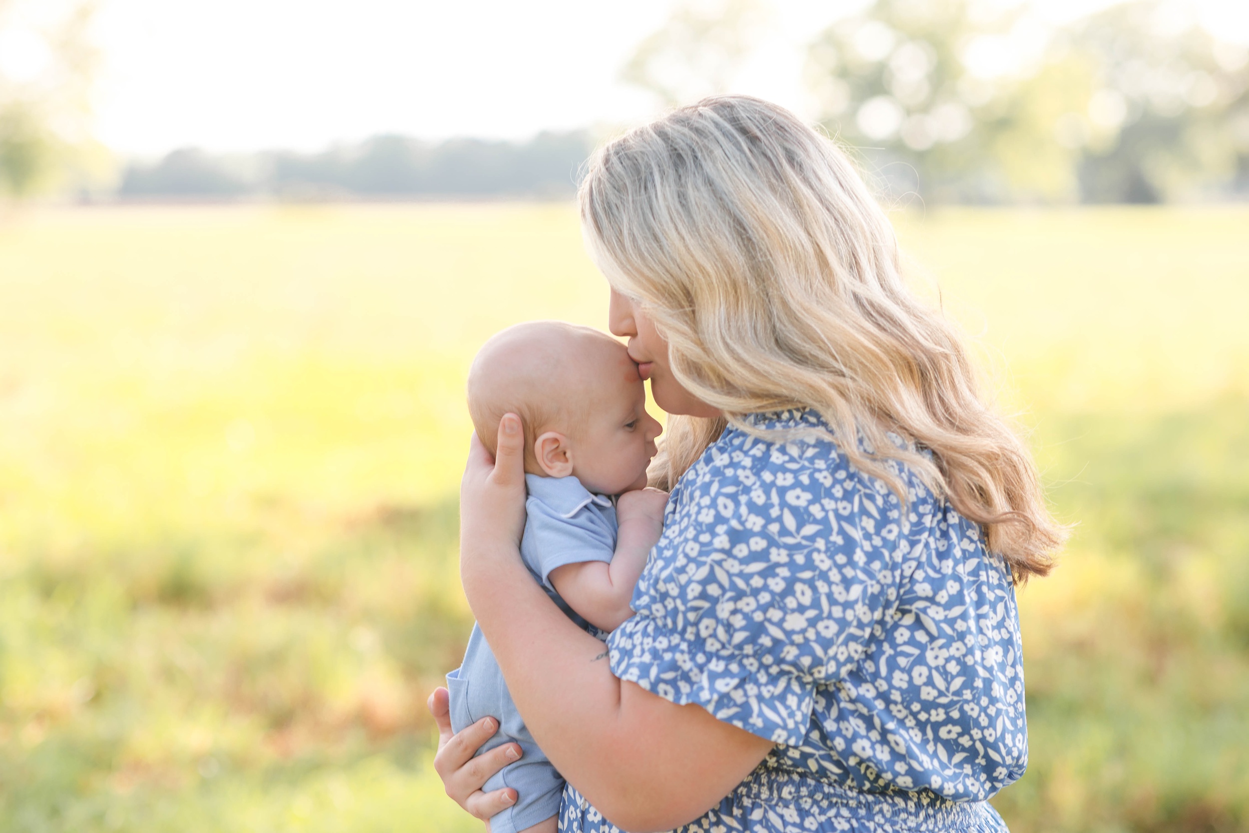 A new mom in a blue dress kisses her newborn baby's head while standing in a field after visiting a toy store in savannah