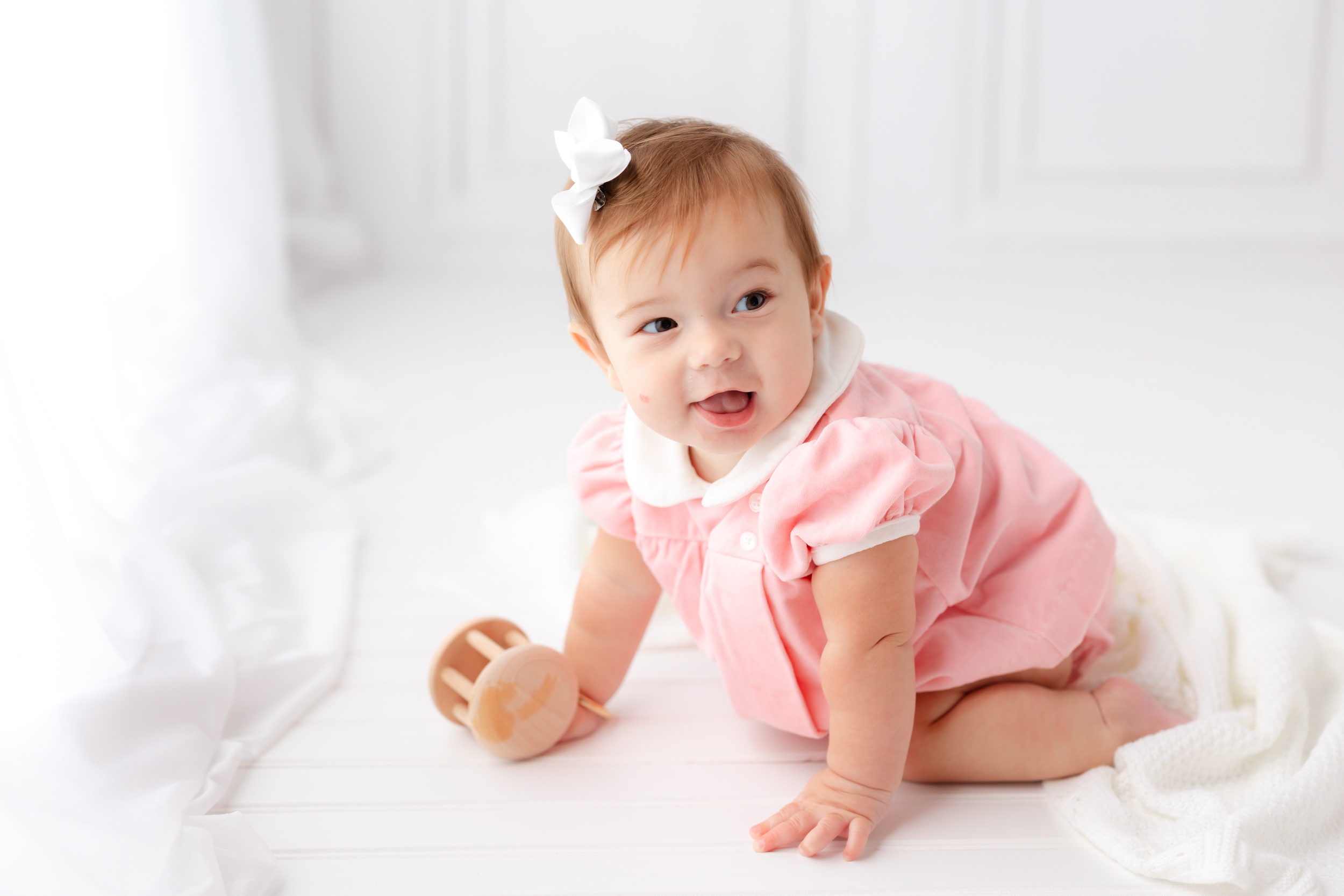 A happy baby in a pink onesie and white bow plays with wooden toys in a studio