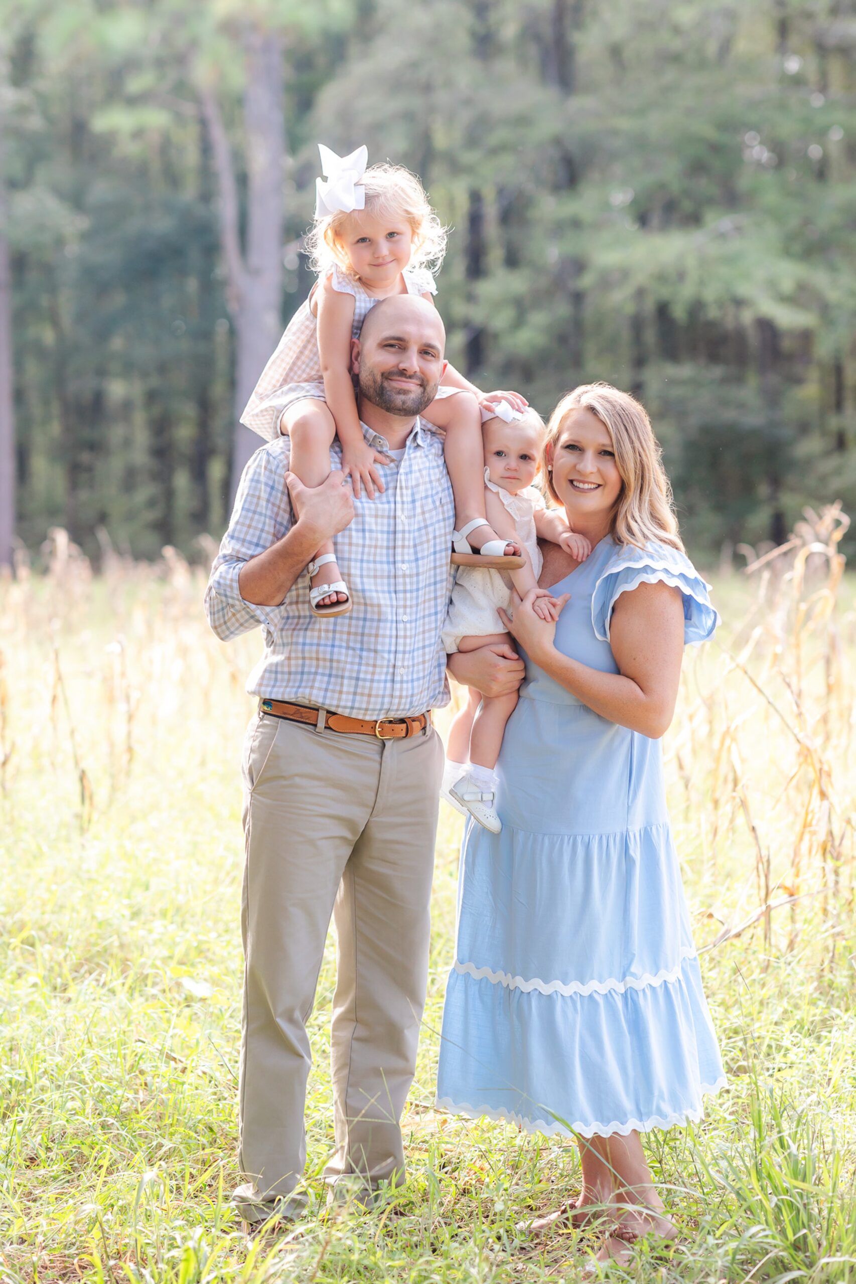 A smiling mom and dad stand in a field of tall grass with their two toddler daughters on their shoulder's and hip before visiting Children's Museum of Pooler