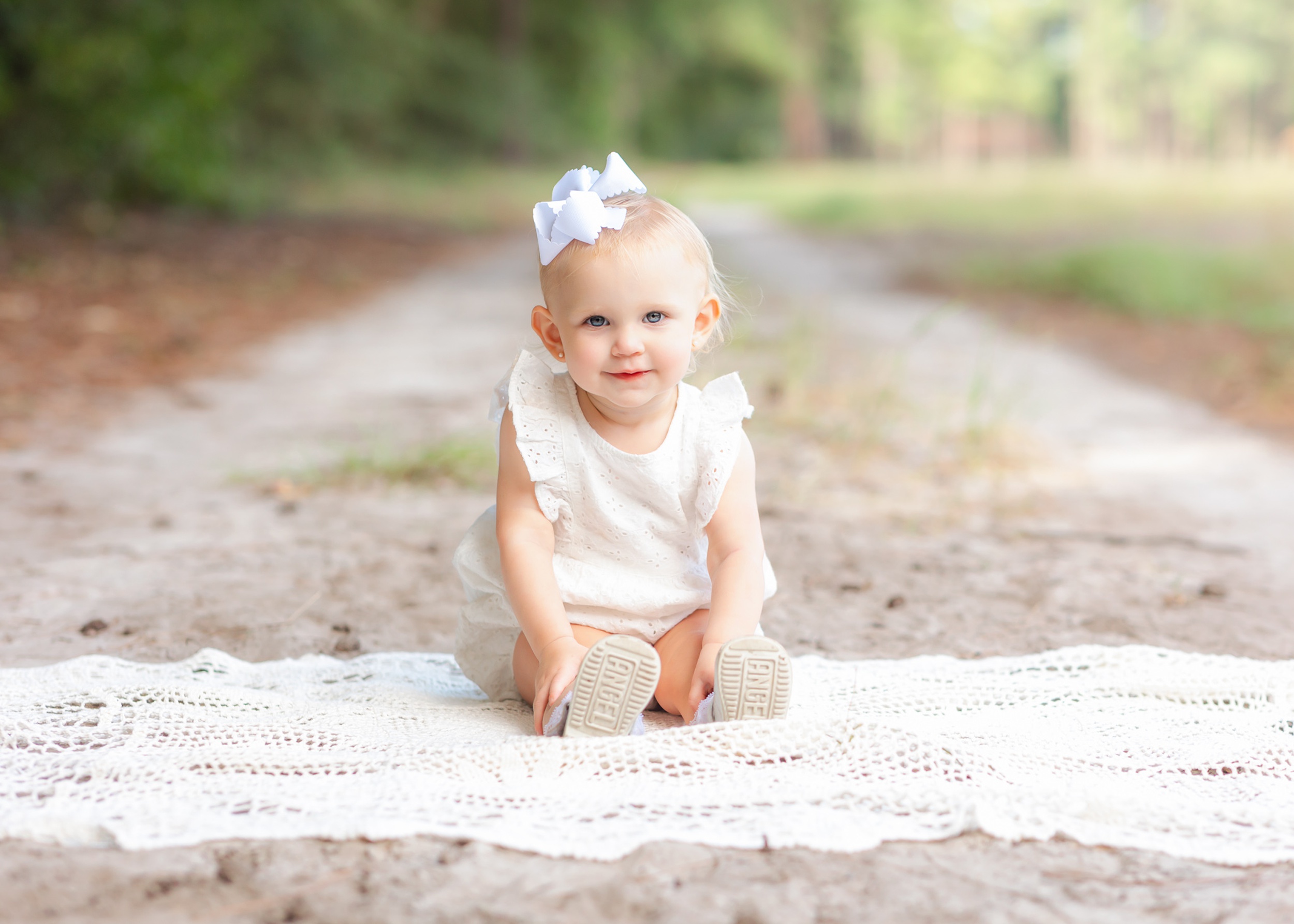 A baby girl in a white onesie sits on a lace blanket in a park trail before exploring Children's Museum of Pooler