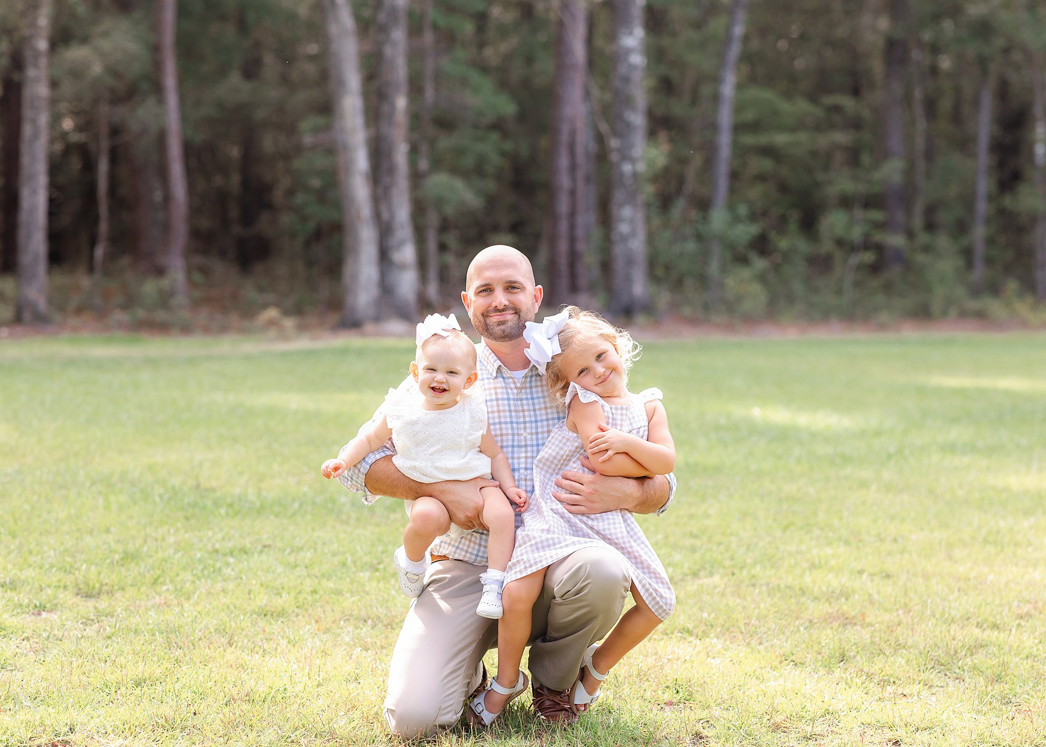 A proud dad kneels in a lawn with her toddler daughters in his arms