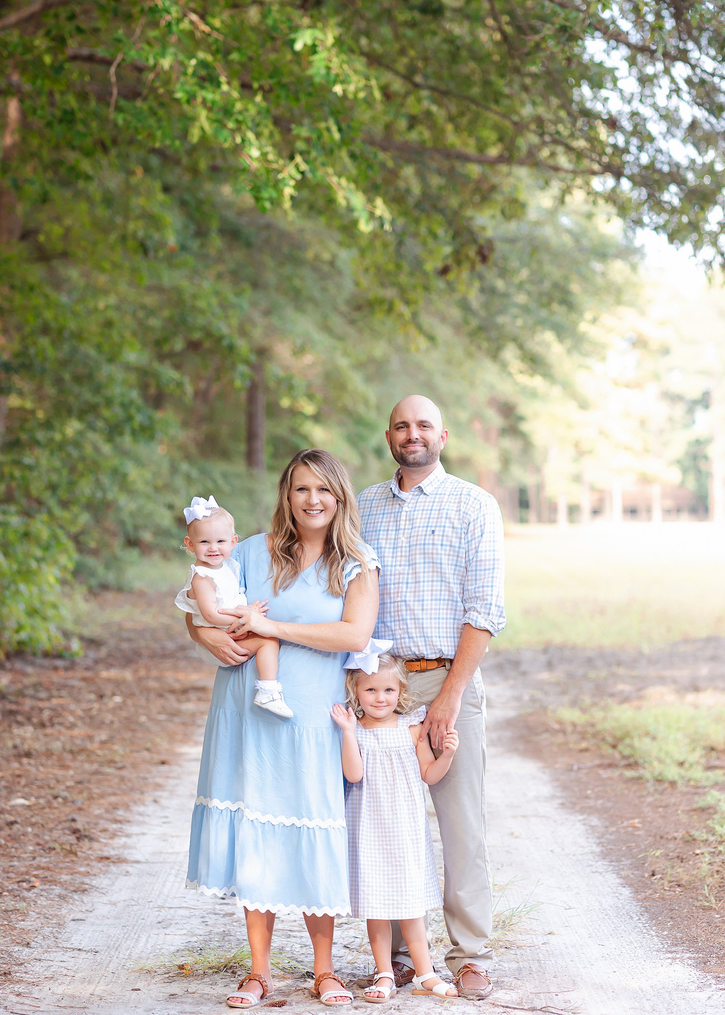 A happy family of 4 with 2 toddler girls stands in a park trail holding hands while finding Family friendly places to stay in Savannah, GA