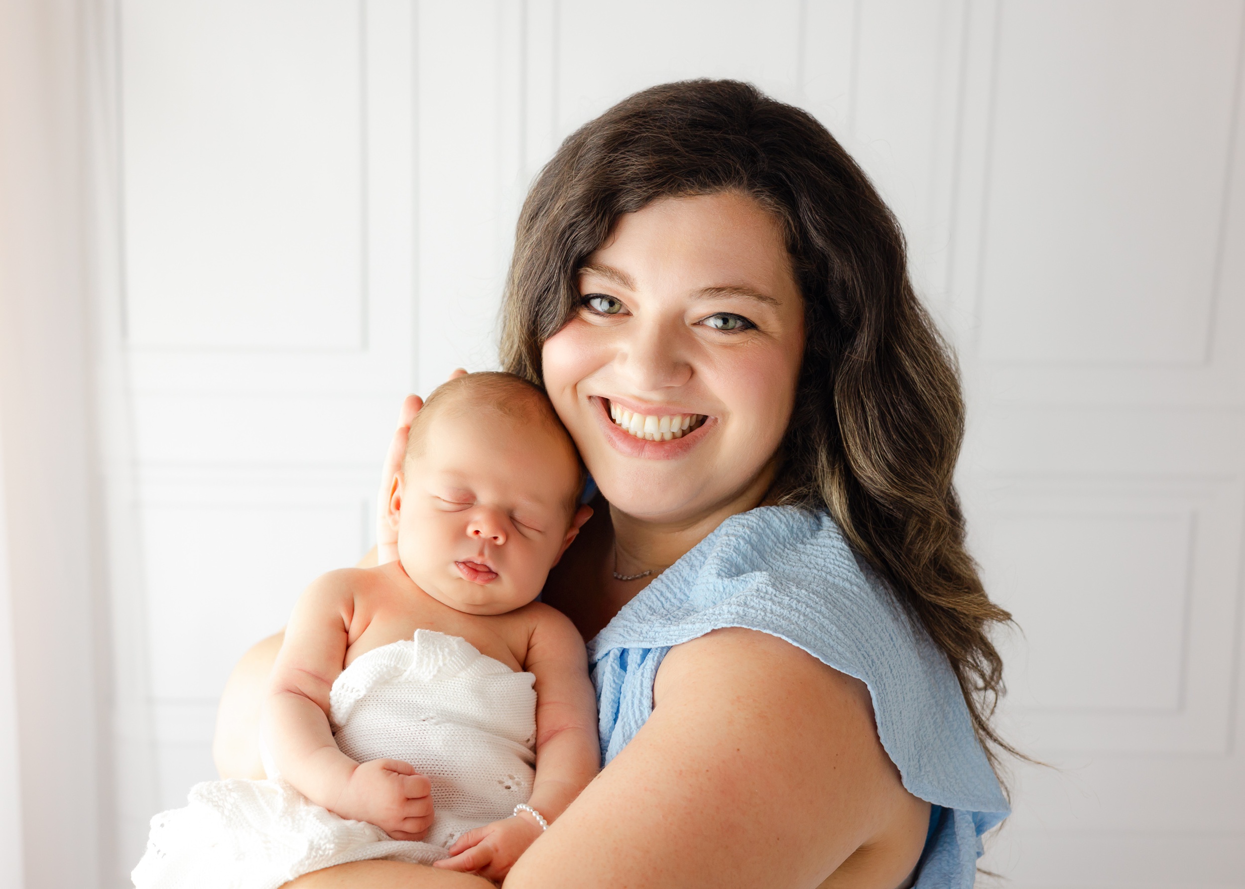 A happy new mom cradles her sleeping newborn against her cheek while smiling big