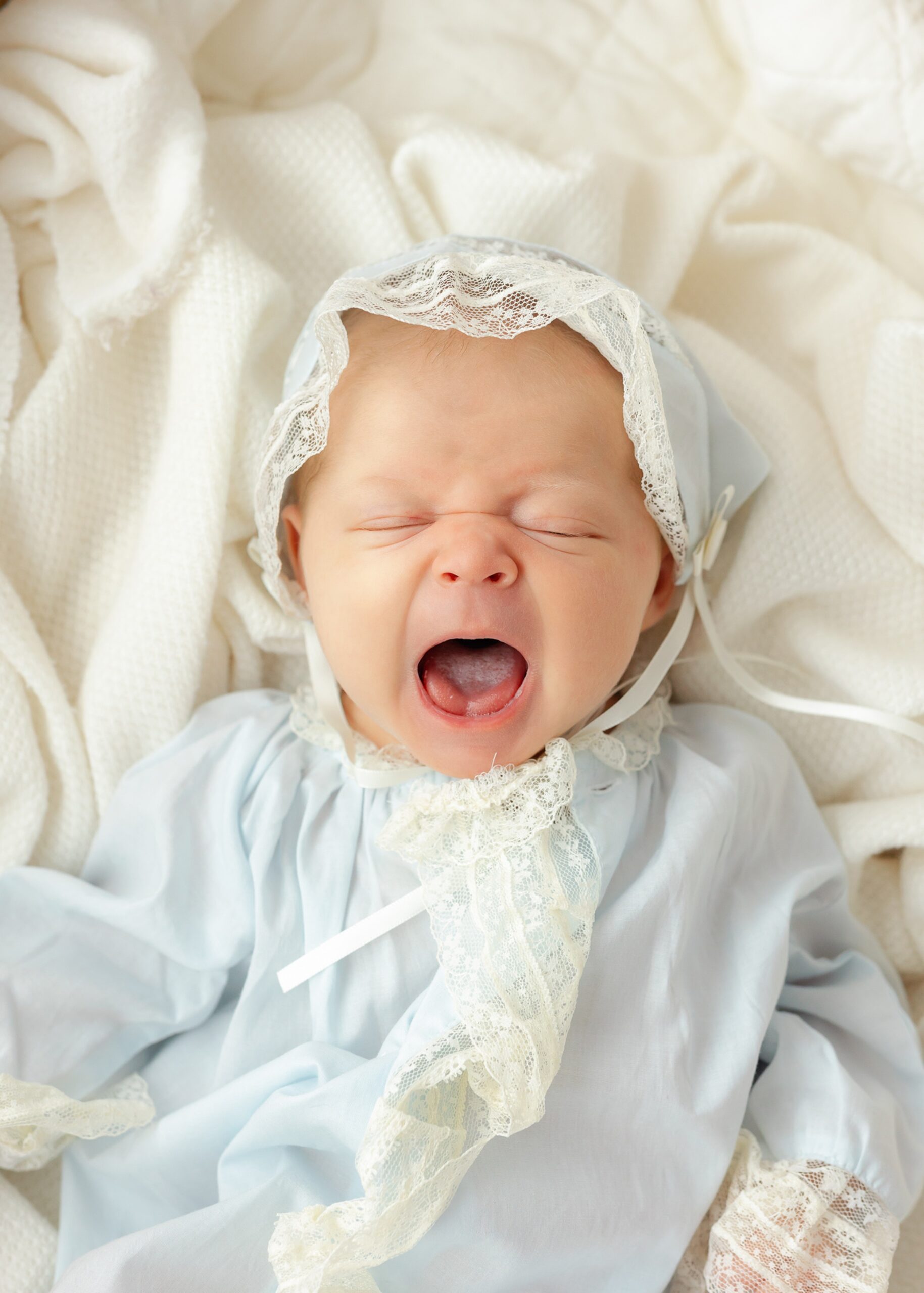 A newborn baby in a blue bonnet and matching onesie yawns in a bed after visiting Pink Chicken Savannah