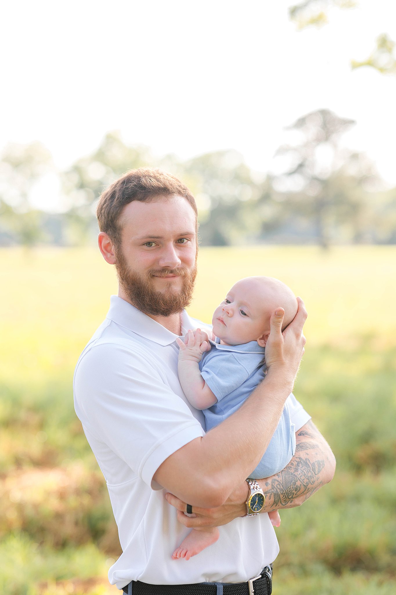 A happy dad holds his baby son in a lawn at sunset in a whtie polo after visiting pools in Savannah, GA