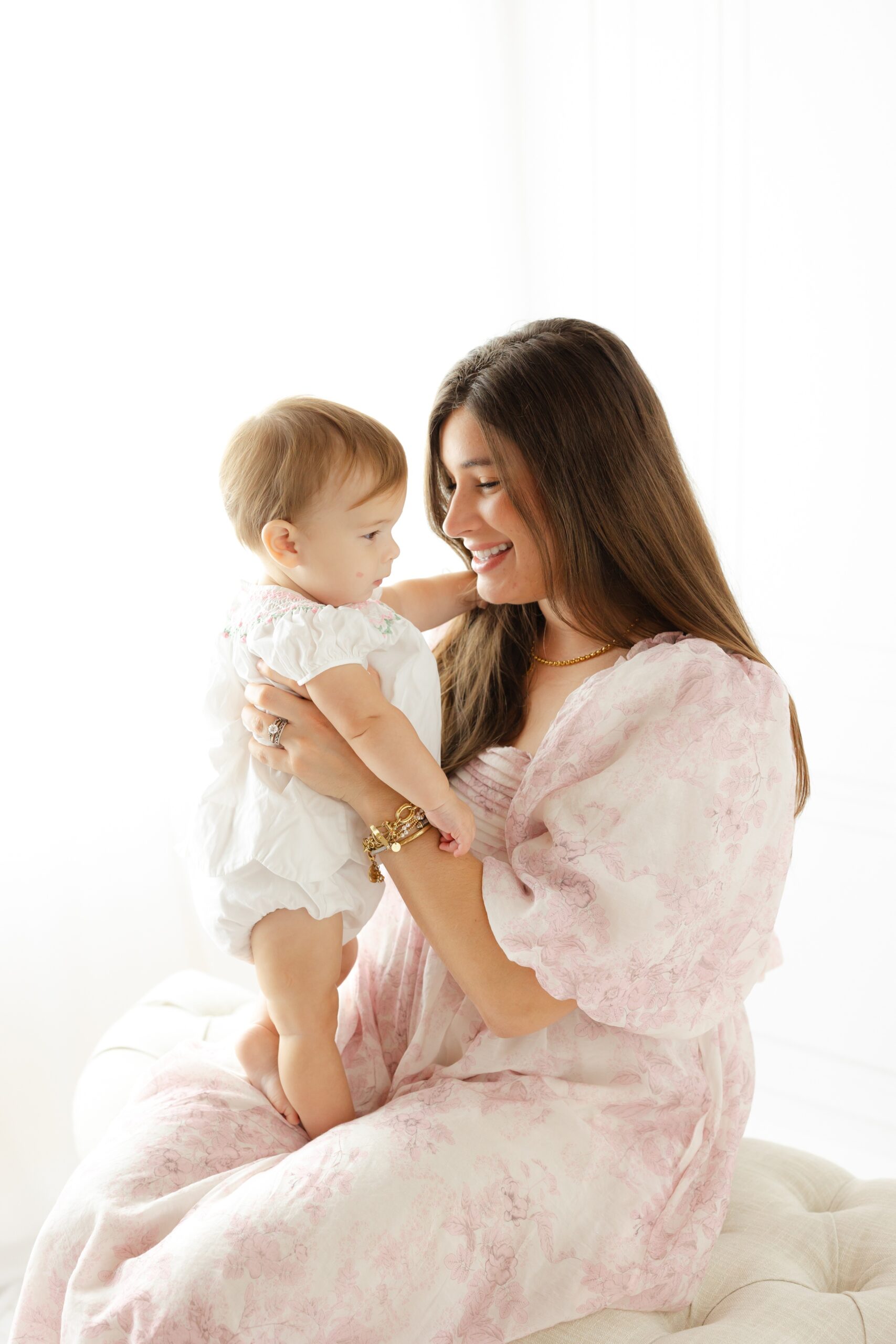 A happy mom sits on a bench in a studio playing with her baby in a white and pink dress before some things to do with kids in savannah