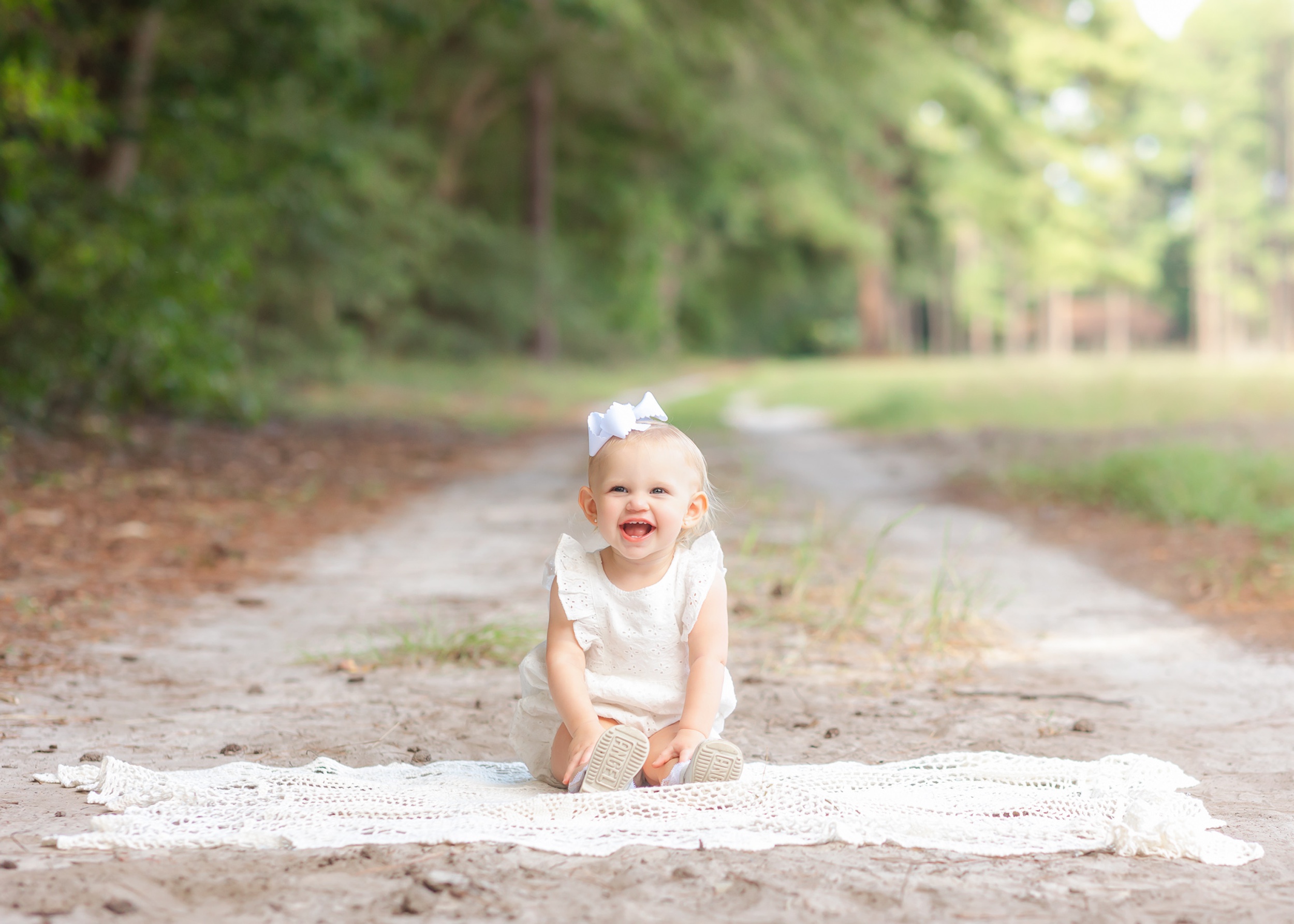 A happy toddler girl in a white dress sits on a blanket in a park trail