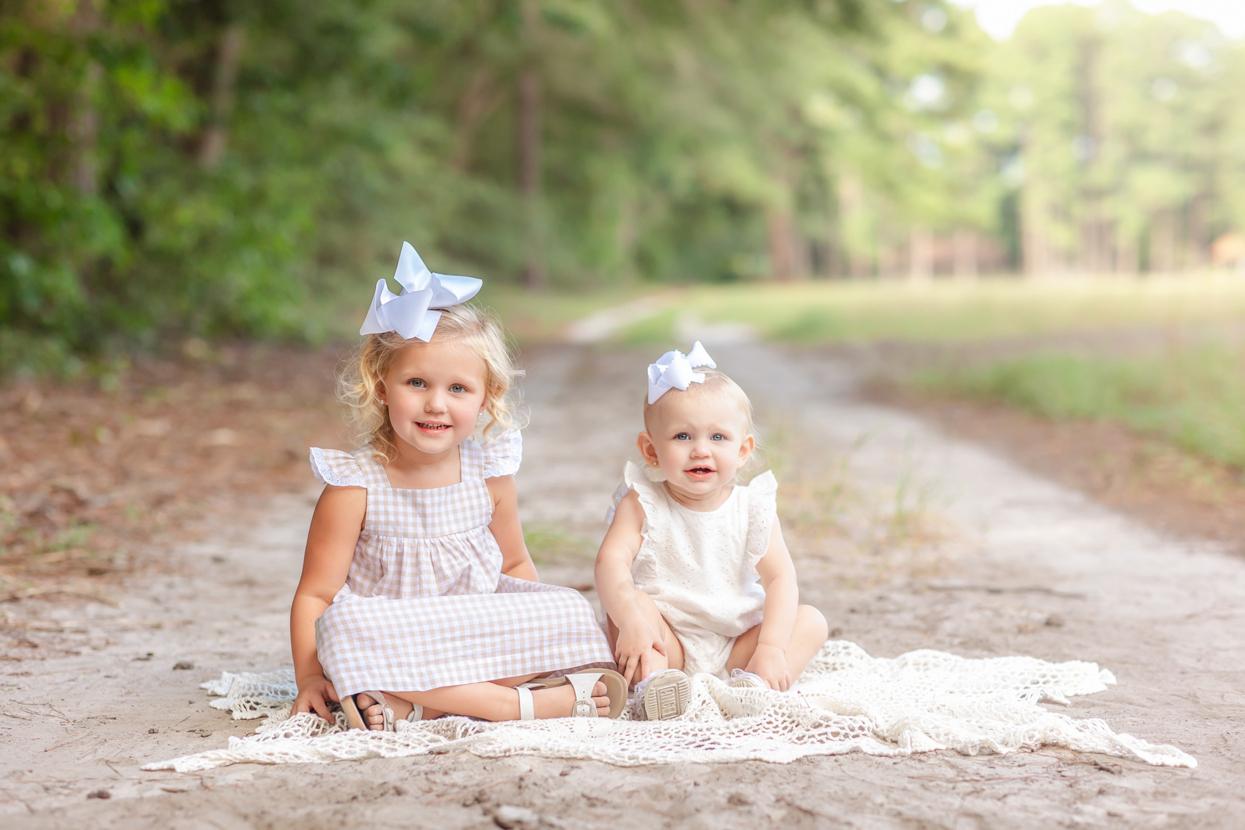 Two baby girls in white dresses sit on a blanket in a park trail smiling before some after school programs in Savannah, GA