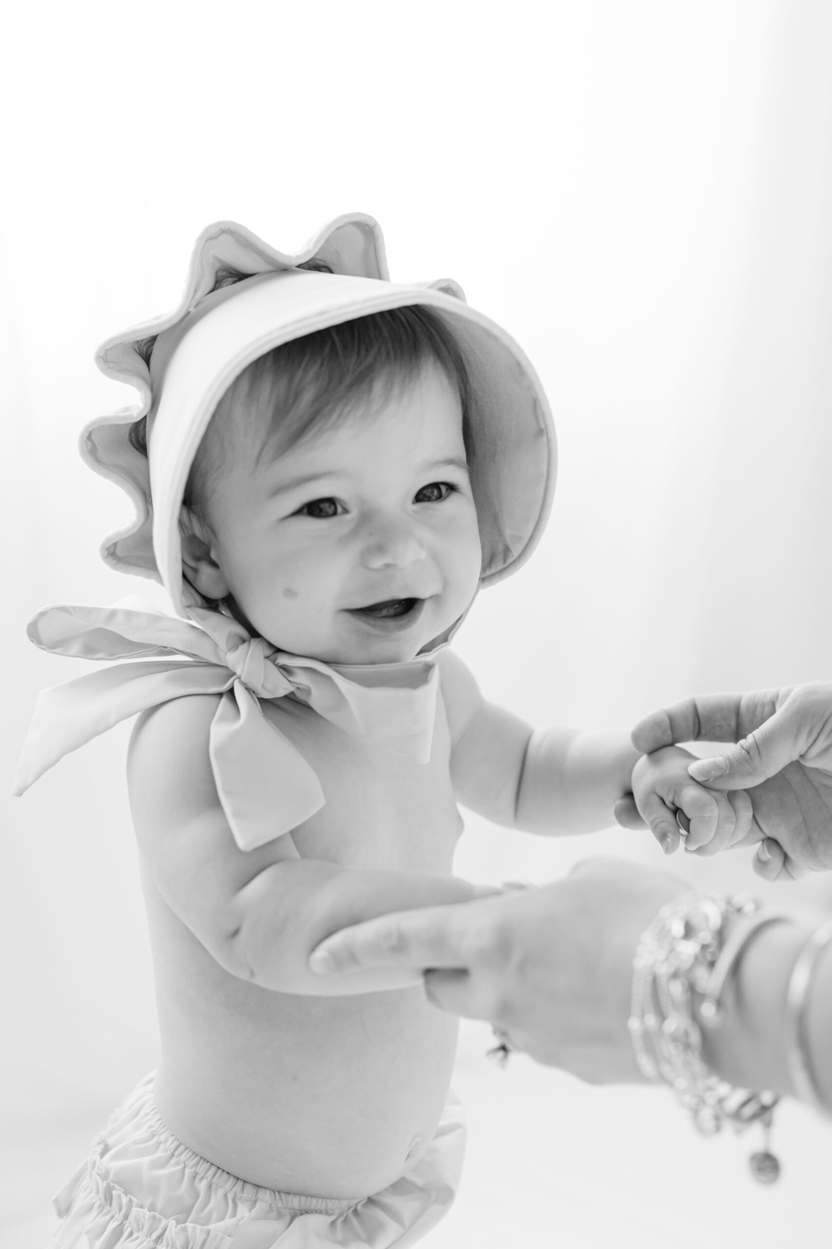 A happy baby in a bonnet walks and smiles in a studio while holding mom's hands before some birthday parties in savannah ga