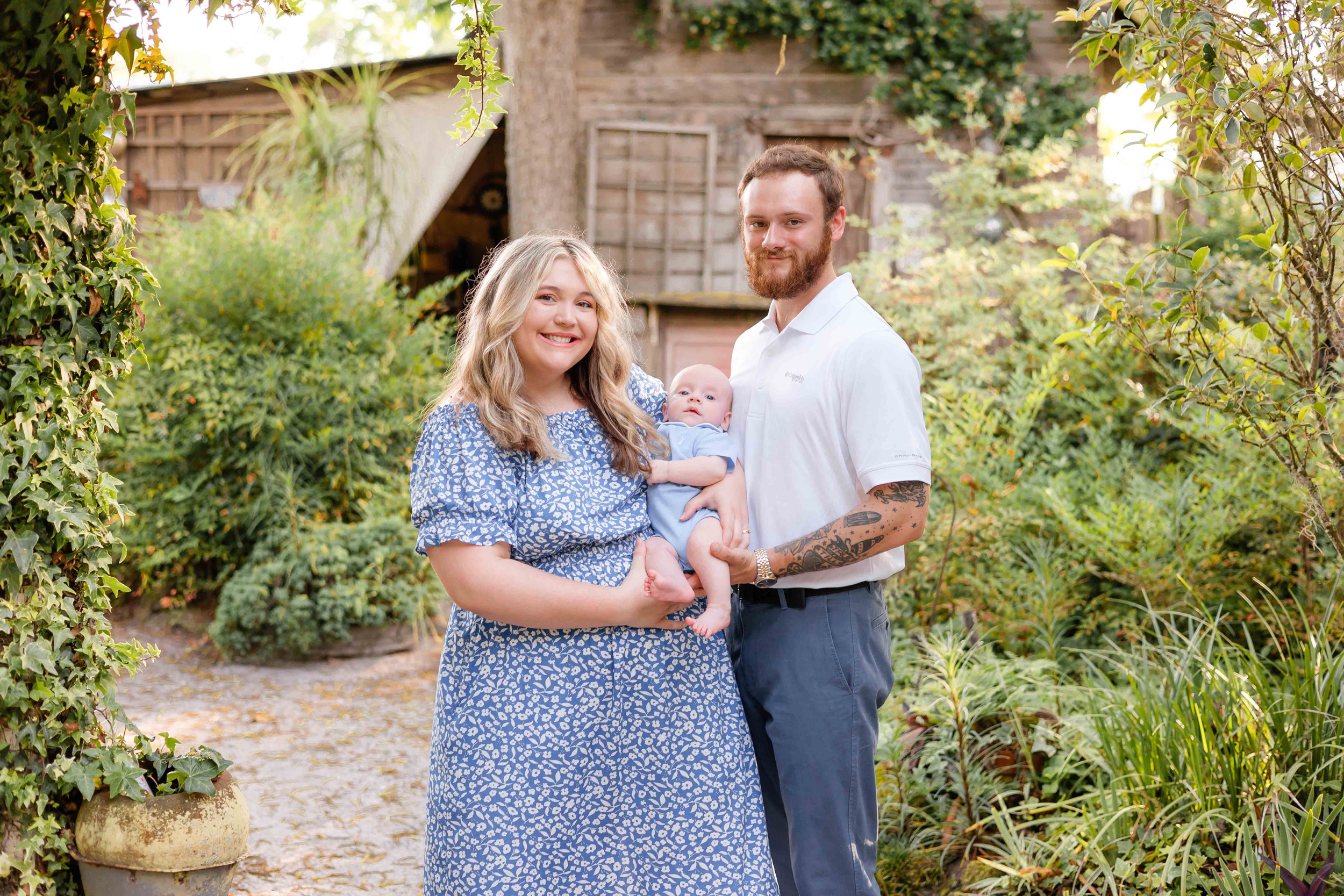 A happy mom and dad in blue stand in a garden cradling their newborn while exploring parks in savannah