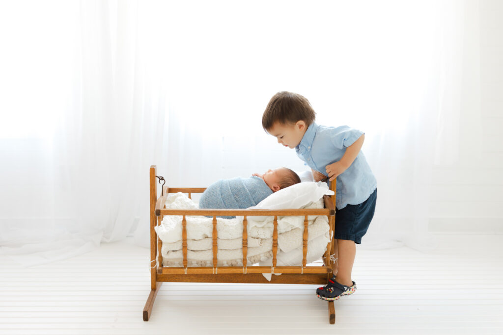 Newborn baby boy wrapped in a blue yarn wrap. Sitting in an antique crib while his big toddler brother looking down at him smiling.