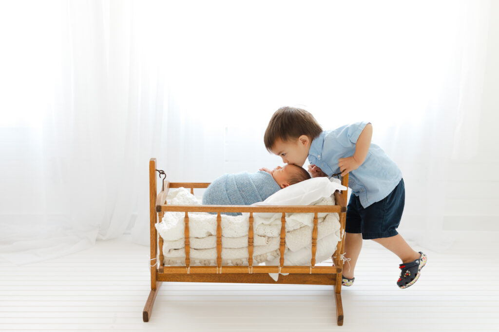 Toddler big brother leaning over an antique brown crib to kiss his newborn brother.