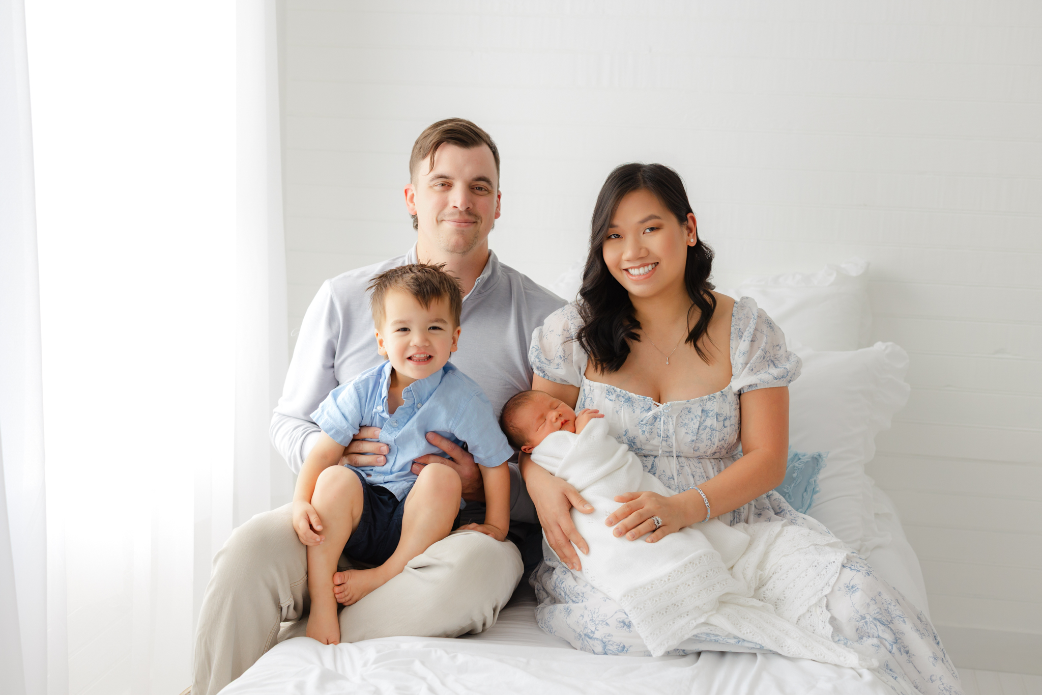 Family of 4 smiling at the camera with their toddler son and newborn baby. Sitting on a white bed.