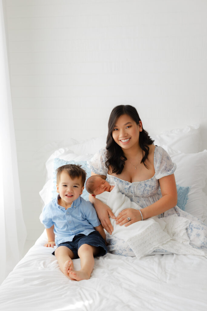 Young mom holding newborn baby boy sitting beside a smiling toddler big brother