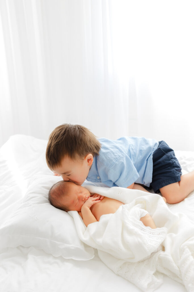 toddler boy kissing his newborn brother on a white bed.