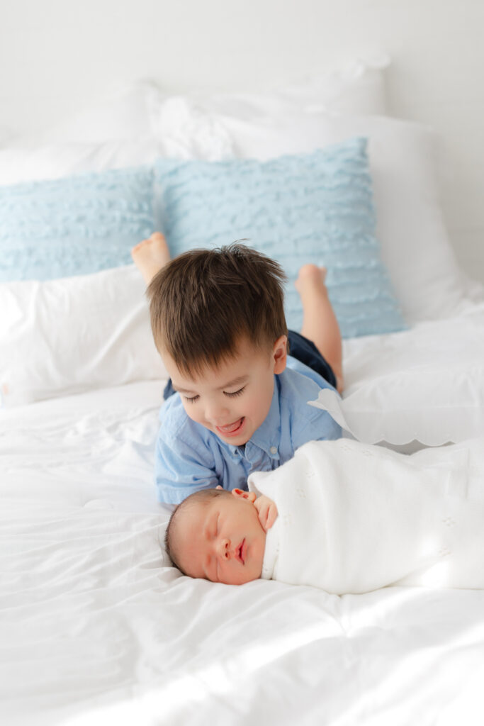 Toddler boy wearing blue smiling at his baby brother wrapped in a white blanket in Savannah, ga