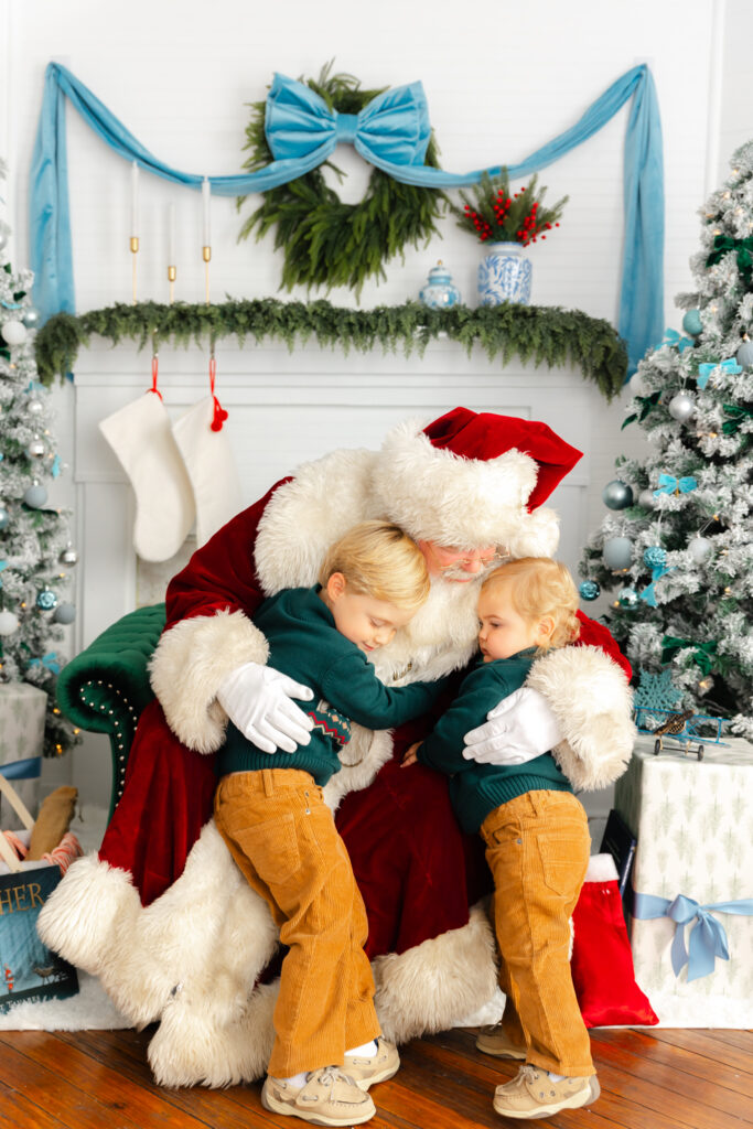two little boys hugging Santa who is sitting in a emerald green chair
