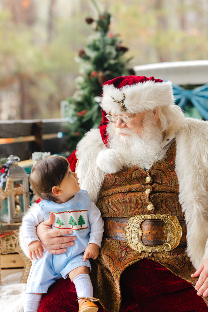 Baby boy in blue holiday outfit looking up at Santa during a private Santa Experience in Savannah, GA.